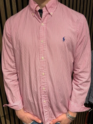 Rödvit randig skjorta från Ralph Lauren - Klassisk röd och vit randig skjorta från Ralph Lauren med blå broderad logga på bröstet. Skjortan har lång ärm, knappar framtill och button-down krage. Tillverkad i bomull och har en normal passform.