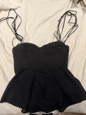 Svart korsettopp med volang - Snygg svart korsettopp med tunna axelband och volang nedtill. Toppen har en figurnära passform och markerad byst, vilket ger en feminin siluett. Perfekt att styla med jeans eller kjol för en trendig look.