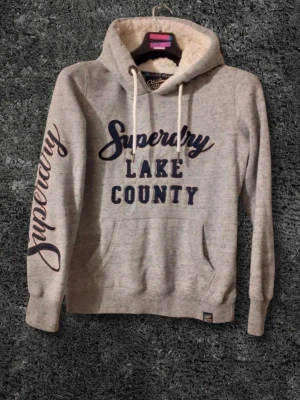 Superdry grå hoodie med tryck - Grå hoodie från Superdry med blått tryck 'Superdry LAKE COUNTY' på bröstet och 'Superdry' längs ena ärmen. Mjuk fuskpäls i huvan, vita dragsnören och känguruficka framtill. Perfekt för dig som gillar streetwear och vill ha något riktigt bekvämt. Storlek 10 sitter som m 