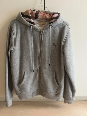 Grå zip hoodie från Burberry - Snygg grå hoodie från Burberry med dragkedja och klassiskt rutigt foder i huvan. Har två fickor framtill, dragsko i huvan och diskret Burberry-logga broderad på bröstet. Tillverkad i mjukt bomullsmaterial för en skön känsla.