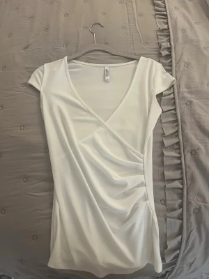 Vit omlottopp med kort ärm - Snygg vit omlottopp med korta ärmar och v-ringning. Toppen har en figurnära passform och är gjord i ett mjukt, stretchigt material som känns bekvämt mot huden. Perfekt att styla med jeans eller kjol för en clean look.