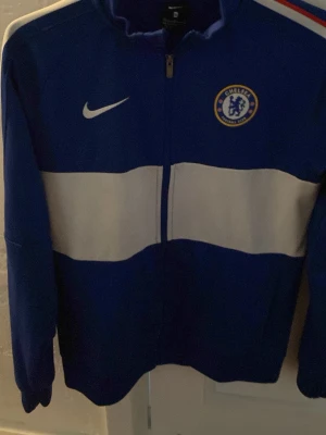 Chelsea Nike blå träningsjacka - Snygg blå träningsjacka från Nike med Chelsea FC-logga på bröstet och vit horisontell rand över magen. Jackan har dragkedja framtill och hög krage. Perfekt för fotbollsträning eller att bära till vardags.        Storlek barn XL