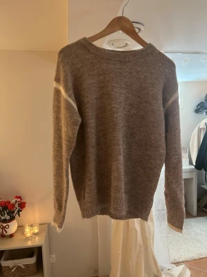 Beige stickad tröja med vit detalj - Mysig beige stickad tröja med rund hals och diskreta vita ränder vid axlarna. Tröjan har en avslappnad passform, ribbstickade muddar och är perfekt för lager-på-lager. Materialet ser ut att vara en mjuk ull- eller ullblandning.