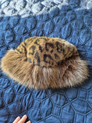 Leopardmönstrad pälsmössa - Säljer en fluffig pälsmössa med leopardmönster i brunt och svart. Mössan har en bred kant i ljusbrun fuskpäls som ger en lyxig och retro vibe. Perfekt för dig som vill sticka ut och hålla värmen under kalla dagar.