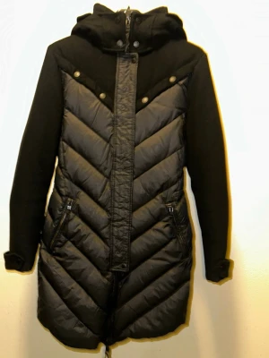 J.LindebergSvart quiltad kappa med huva  - Snygg svart kappa som framhäver de kvinnliga formerna från J.Lindebergmed quiltade partier och huva. Jackan har dragkedja framtill, tryckknappar och två fickor med dragkedja. Materialmix med vadderad front och ärmar i mjukare tyg. Perfekt för kyliga dagar.