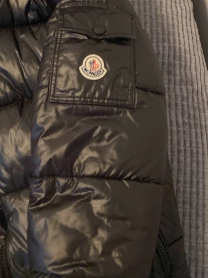 Svart dunjacka från Moncler - Snygg svart dunjacka från Moncler med glansig finish och klassisk logotyp på ärmen. Jackan har en ficka med knapp på ärmen och quiltad design som håller värmen under kalla dagar. Perfekt för dig som vill ha både stil och funktion.