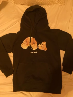 Svart Palm Angels hoodie med nalle - Säljer en svart hoodie med en ikonisk nallebjörn-tryck på bröstet och vit logga under. Hoodien har huva med dragsko och en stor magficka. Perfekt för dig som gillar streetwear och vill sticka ut med en unik design.