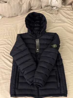  pufferjacka från Stone Island - Snygg  pufferjacka från Stone Island med huva och dragkedja framtill. Jackan har det klassiska Stone Island-märket på vänster ärm.