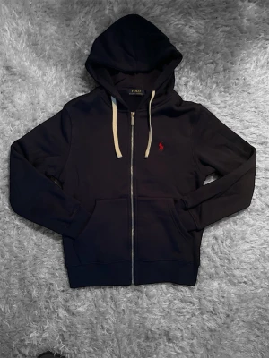 Marinblå hoodie från Polo Ralph Lauren - Snygg marinblå hoodie från Polo Ralph Lauren med dragkedja, vit dragsko och röd logga broderad på bröstet. Klassisk passform med två fickor framtill och mjukt material som känns skönt mot huden. Perfekt för en avslappnad och stilren look.