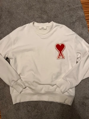 Vit sweatshirt från Ami Paris - Säljer en vit sweatshirt från Ami Paris med det ikoniska röda hjärtat och A-loggan broderad på bröstet. Tröjan har rund hals, ribbade muddar och är gjord i mjuk bomull. Perfekt för en clean och trendig look. Vet ej om den är äkta därför det billiga priset!!