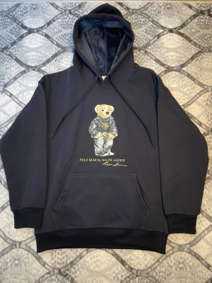 Svart hoodie Polo Bear Ralph Lauren - Snygg svart hoodie från Ralph Lauren med Polo Bear-tryck på bröstet. Klassisk känguruficka framtill, ribbade muddar och justerbar huva. Mjuk insida och cool björn i kostym ger en unik touch. Perfekt för dig som vill sticka ut med stilren streetwear.
