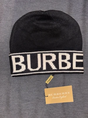 Svart mössa från Burberry - Snygg svart stickad mössa från Burberry med vit logotyp i stora bokstäver runt kanten. Klassisk och enkel design som passar till det mesta. Tillverkad i mjukt material för skön känsla och stilren look.