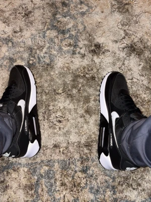 Nike Air Max 90 svartvita sneakers - Säljer ett par Nike Air Max 90 sneakers i svart och vitt. Skorna har en klassisk låg siluett, vit sula med synlig Air Max-dämpning och svart ovandel i mesh och läder. Ikonisk Swoosh-logga på sidan och detaljer i vitt och svart.