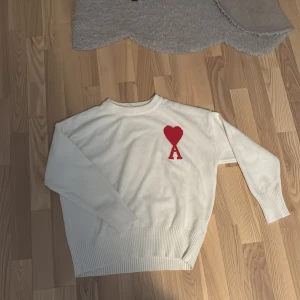 Vit stickad tröja från Ami Paris - Vit stickad tröja från Ami Paris med röd hjärtlogga och bokstaven A på bröstet. Tröjan har rund hals, ribbade muddar och lång ärm. Perfekt för dig som gillar clean stil med en twist av statement-detalj. Den är i storlek S men passar för de som har storlek M.