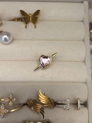 Hjärthänge med guldtonad ram - Sött smyckeshänge i form av ett hjärta med en gnistrande, klar sten i mitten och guldfärgad ram. Hänget har en pil genom hjärtat vilket ger en romantisk vibe. Perfekt att använda som berlock till halsband eller armband.