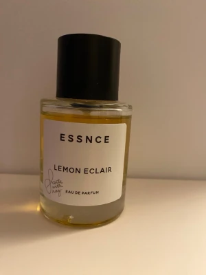 ESSNCE Lemon Eclair Eau de Parfum - Fräsch parfym från ESSNCE med doften Lemon Eclair. Kommer i en rund glasflaska med svart lock och vit etikett. Parfymen har en gulaktig vätska och modern, minimalistisk design. Perfekt för dig som gillar citrusdofter och stilrena flaskor. Limited edition. Tillverkas inte mer. Mängd enl bilder 