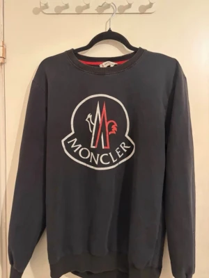 Svart Moncler sweatshirt med logga - Cool svart sweatshirt från Moncler med stor vit och röd logga framtill. Tröjan har rund halsringning, ribbade muddar och är tillverkad i mjukt material. Perfekt för en avslappnad och trendig stil.