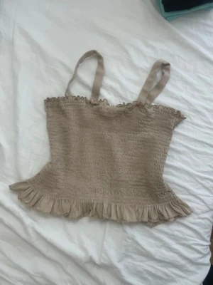 Beige linnetopp med volangdetalj - Supersöt beige linnetopp med smockad resår, volangkant nertill och justerbara axelband med knapp. Perfekt för varma dagar och enkel att styla med jeans eller kjol.