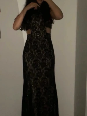 Svart långklänning med spetsdetaljer - Elegant svart långklänning med spetsdetaljer och glittriga inslag. Klänningen har ärmlös design, markerad midja med öppna partier i sidorna och figurnära passform. Perfekt för speciella tillfällen där du vill sticka ut.