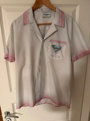 Casablanca Tennis Club skjorta - Säljer en vit kortärmad skjorta från Casablanca med rosa detaljer och tryck på bröstficka och rygg. Skjortan har klassisk krage och motiv med palmer, tennisbana och texten 'Tennis Club'. Perfekt för dig som gillar sportig och trendig stil.