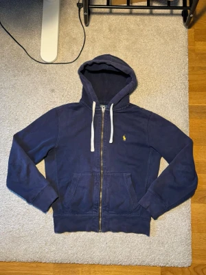 Marinblå zip från Polo Ralph Lauren (Äkta) - (Äkta) Mörkblå zip med dragkedja från Polo Ralph Lauren. Klassisk design med vit dragsko i huvan, kängurufickor och gul broderad logga på bröstet. Tillverkad i mjuk bomullsblandning för skön känsla.