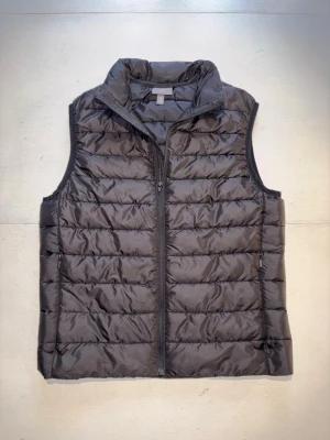 Svart dunväst från H&M - Snygg svart dunväst med quiltad design och ståkrage. Västen har dragkedja framtill och två sidofickor med dragkedja. Perfekt för lager-på-lager och ger en clean look till din outfit. Materialet är lätt och håller värmen utan att kännas klumpig. Storlek S. Skick 9/10.