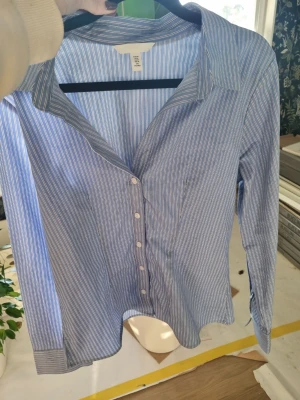 Blåvit randig skjorta från H&M - Klassisk blå skjorta med vita ränder från H&M. Skjortan har lång ärm, krage och knäppning framtill med vita knappar. Tillverkad i bomull för en skön och luftig känsla. Perfekt för en clean och stilren look.