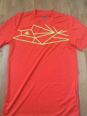 Röd Adidas t-shirt med grafiskt tryck - Snygg röd t-shirt från Adidas med ett gult grafiskt mönster över bröstet. Klassisk rund halsringning och korta ärmar. Perfekt för dig som gillar sportig stil och vill sticka ut lite extra.