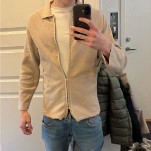 Beige ziptröja med krage från H&M - Snygg beige ziptröja från H&M med klassisk krage och lång ärm. Tillverkad i ett mjukt, lätt material som ger en avslappnad look. Perfekt att bära över en t-shirt eller som lager. Enkel design utan synliga loggor eller mönster.