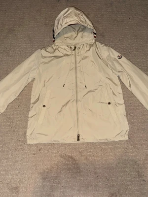 Beige vindjacka från Moncler - Säljer en beige vindjacka från Moncler med huva och dragsko. Jackan har hel dragkedja framtill, två fickor med knapp och Moncler-logga på ärmen. Huvan har snygga ränder i rött, vitt och blått. Perfekt för vår och höst när du vill ha en clean och stilren look.