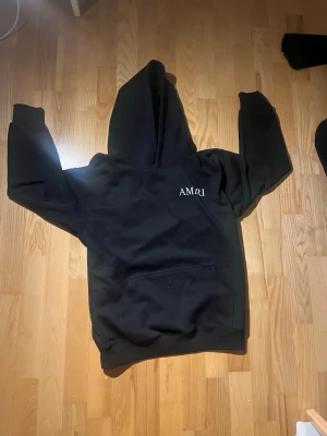 Svart hoodie från Amiri med tryck - Säljer en svart hoodie från Amiri med vit logga på bröstet och stort tryck med text och motiv på ryggen. Hoodien har klassisk känguruficka och stor huva. Perfekt för dig som gillar streetwear och vill sticka ut med snygga detaljer.