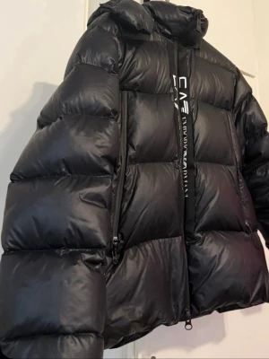 Svart pufferjacka från EA7 Emporio Armani - Säljer en svart pufferjacka från EA7 Emporio Armani med stor huva och feta vadderade paneler. Jackan har dragkedja framtill, två sidofickor med dragkedja och EA7-logga på bröstet samt ärmen. Materialet är glansigt och ger en riktigt modern vibe.