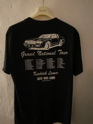Kendrick Lamar Grand National Tour T-shirt - Svart t-shirt med vit tryck fram och bak från Kendrick Lamars Grand National Tour. Framsidan har texten 'GLORIA AUTO BODY' och baksidan visar en bil och turnédatum. Klassisk rund hals och korta ärmar. Perfekt för fans av Kendrick Lamar.