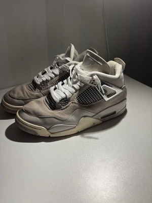 Nike Air Jordan 4 - Snygga Nike Air Jordan 4 Retro i vita och grå toner. Skorna har meshpaneler, mockadetaljer och vita snören. Ikonisk siluett med synlig Air-sula och coola plastdetaljer på sidorna. Perfekta för dig som gillar streetwear och klassisk sneakerstil.