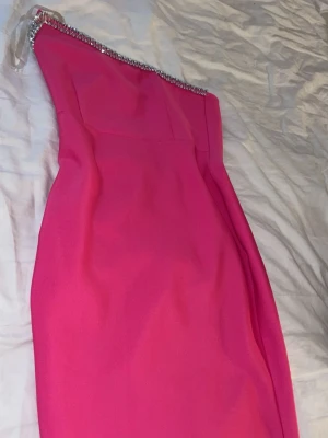 Rosa one shoulder klänning med strass - Snygg rosa klänning med one shoulder-design och strassdetaljer längs axeln. Klänningen har en figurnära passform och är perfekt för dig som vill sticka ut. Materialet känns mjukt och följsamt. Passar perfekt till en bröllopsklänning mm. 