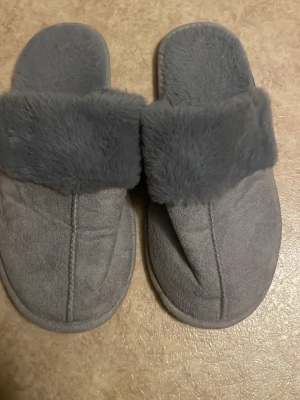 Grå fluffiga tofflor med fuskpäls - Mysiga grå tofflor med mjuk fuskpäls runt kanten och insida. Tofflorna har öppen häl och är tillverkade i ett lent, mockaliknande material. Perfekta för att hålla fötterna varma hemma. Klassisk och enkel design utan synligt märke.