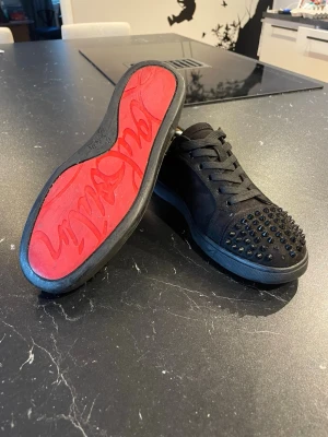 Svarta Louboutin sneakers med nitar - Säljer ett par svarta sneakers från Christian Louboutin med mattsvarta nitar på tån och klassisk röd sula. Skorna har snörning, svart mockaliknande material och cool streetstil. Perfekta för dig som vill sticka ut med en edgy look.