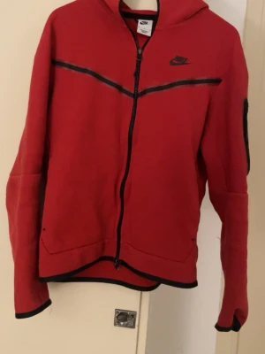 Röd Nike tech - Snygg röd hoodie från Nike med hel dragkedja framtill och svarta detaljer längs bröstet och ärmsluten. Hoodien har en klassisk Nike-logga på bröstet och praktiska fickor. Perfekt för dig som gillar sportig och avslappnad stil.