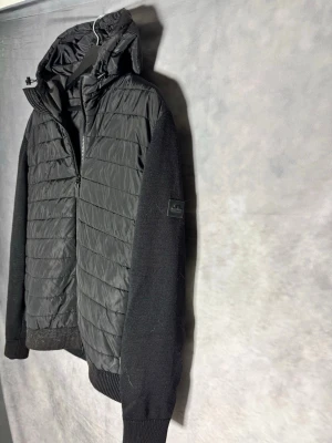 J.Lindeberg - Snygg svart pufferjacka från J.Lindeberg med quiltad front och rygg, stickade ärmar och muddar. Jackan har huva och dragkedja framtill. Perfekt för kalla vinterdagar och ger en modern look. STORLEK L SITTER SOM M