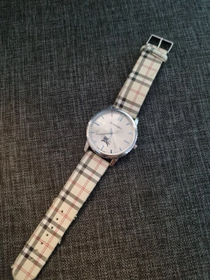 Rutig klocka från Burberry - Stilren klocka från Burberry med klassiskt rutigt armband i beige, svart och rött. Urtavlan är silverfärgad med minimalistiska markeringar och Burberry-logga. Boetten är rund och i metall, vilket ger en tidlös och elegant look.