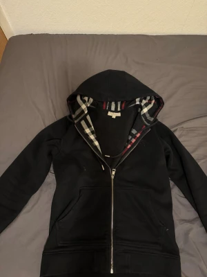 Burberry hoodie med dragkedja - Säljer en svart hoodie från Burberry med klassiskt rutigt foder i huvan och hel dragkedja framtill. Hoodien har två fickor och ribbade muddar. Storlek M och säljer pågrund av att den är för liten, använd fåtal gånger
