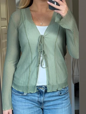 Ljusgrön ribbad mesh blus med knyt - Trendig ljusgrön blus i mesh med ribbad struktur och två knyt framtill. Blusen är långärmad och har en lätt genomskinlig look som ger en cool layering-effekt. Perfekt att styla över ett linne🫶🏼