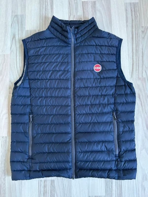 Mörkblå dunväst från Colmar - Snygg mörkblå dunväst från Colmar med quiltad design och hög krage. Västen har två dragkedjeförsedda fickor framtill och Colmar-logga på bröstet. Perfekt att slänga över en hoodie eller tröja för extra värme. Materialet är lätt och smidigt.