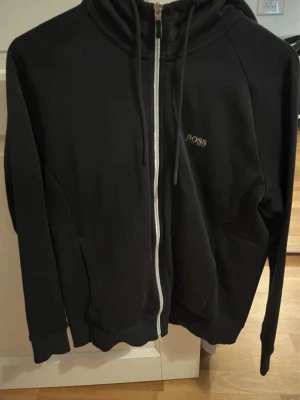 Svart hoodie med dragkedja från BOSS - Snygg svart hoodie från BOSS med vit dragkedja framtill och justerbar huva. Diskret BOSS-logga på bröstet och även baktill. Tillverkad i mjukt material, perfekt för en chill och stilren look. Fickor på sidorna för extra funktion.