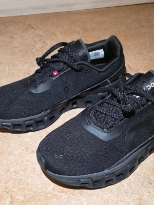 Svarta On Cloud sneakers - Stilrena svarta On Cloud sneakers med ventilerande mesh-ovandel och unika CloudTec-sula för extra komfort. Skorna har svarta snören och diskreta loggor på sidan och tungan. Perfekta för dig som gillar sportig och modern stil.