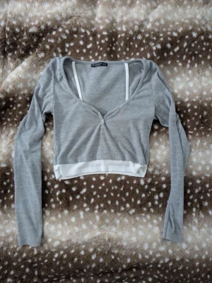 Grå croppad ribbad topp - Grå croppad topp med lång ärm och v-ringning. Toppen har ribbad struktur, vit kant nedtill och knappdetalj framtill. Perfekt för en trendig och avslappnad stil.