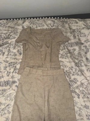 Beige tvådelat set med topp och byxa - Snyggt tvådelat set i beige med kortärmad topp och matchande byxor. Toppen har knappar framtill och en fyrkantig ringning, medan byxorna har raka ben och hög midja 