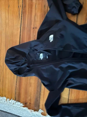 Svart vindjacka från The North Face - Snygg svart vindjacka från The North Face med huva och dragkedja framtill. Jackan har logga på bröstet och ärmen, samt elastiska muddar vid handlederna. Tillverkad i lätt och tåligt material som skyddar mot vind.