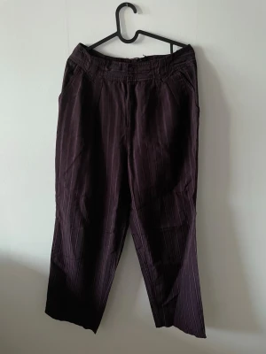 Mörklila randiga kostymbyxor - Mörklila byxor med diskreta vertikala ränder. Byxorna har hög midja, bälteshällor och sidofickor. Materialet känns mjukt och har en lätt glans. Dock tydligt slitage vid byxlinningen.