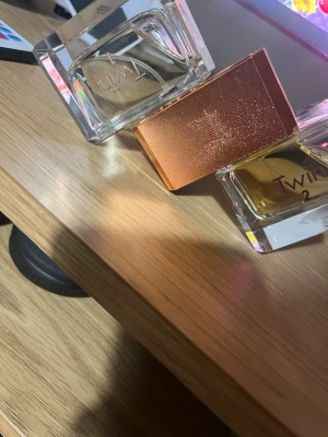 Twin parfym duo med kopparlock - Två stilrena parfymflaskor från Twin med rektangulär form och transparent glas. Ena flaskan har ett elegant kopparfärgat lock med stjärnmönster. Båda flaskorna har guldfärgad vätska och minimalistisk design, perfekt för dig som gillar moderna dofter.